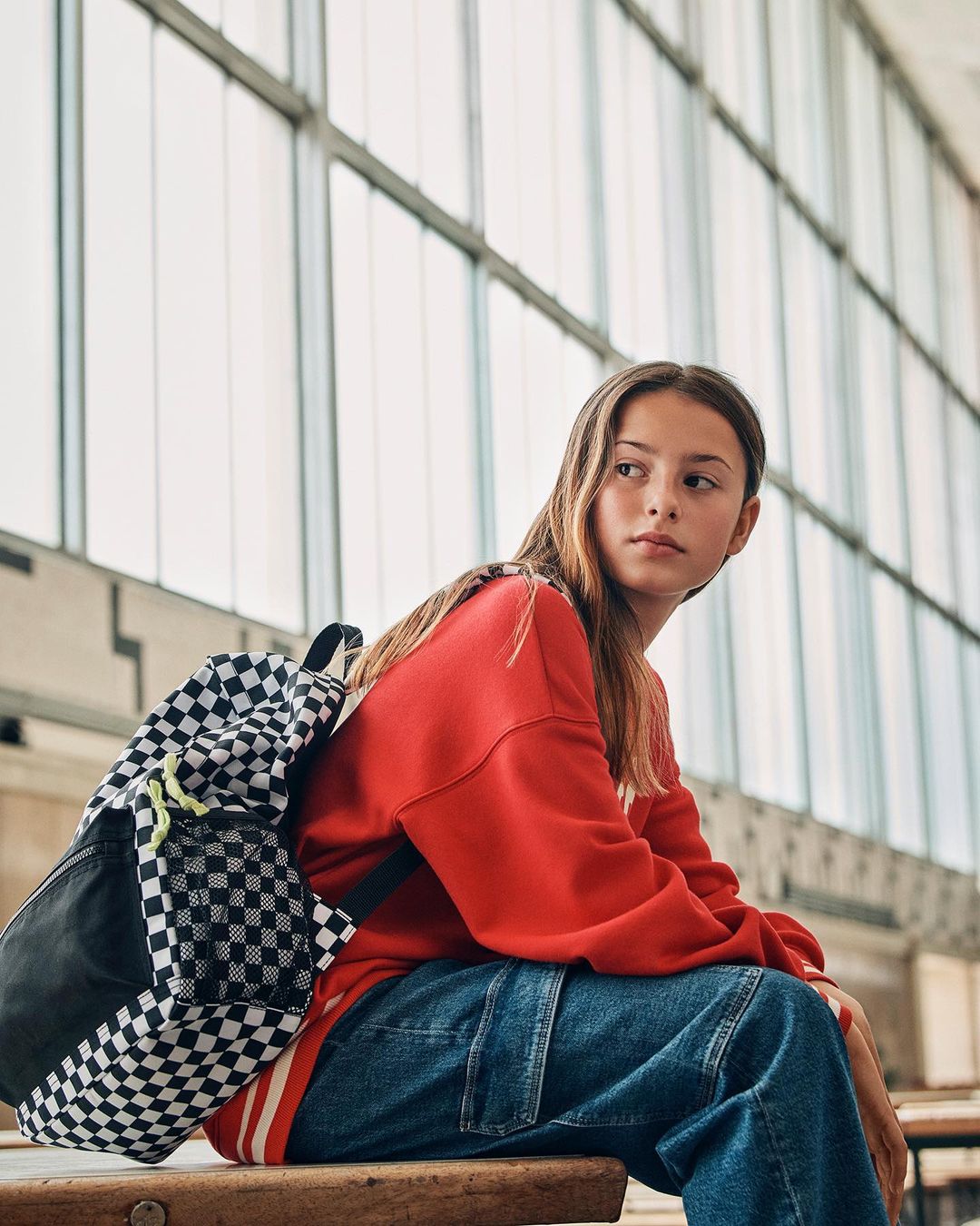 Teenager poses indoors wearing red top and jeans with backpack. Large windows behind provide diffused natural lighting.