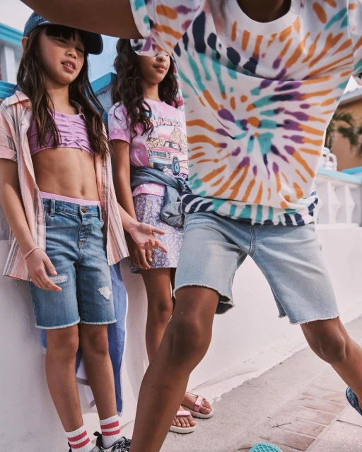 Three young girls pose outdoors wearing casual summer clothing, including denim shorts, a tie-dye shirt, and a baseball cap.