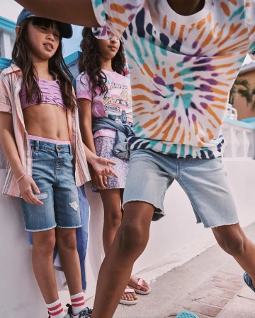 Three young girls pose outdoors wearing casual summer clothing, including denim shorts, a tie-dye shirt, and a baseball cap.