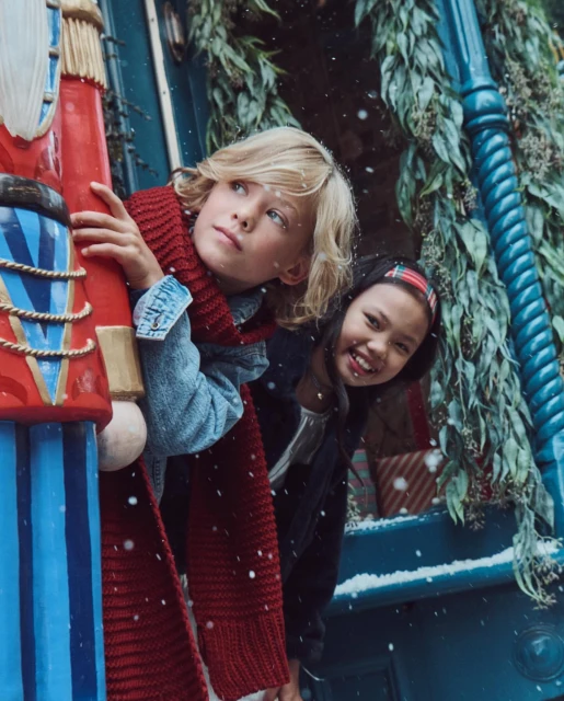 Two kids, a boy and a girl, pose near a large toy nutcracker with gifts and greenery, snow falling gently, evoking a holiday atmosphere.
