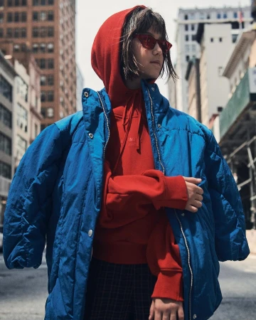 Woman in blue puffer jacket and red hoodie stands in an urban setting, featuring tall buildings in the background on a sunny day.