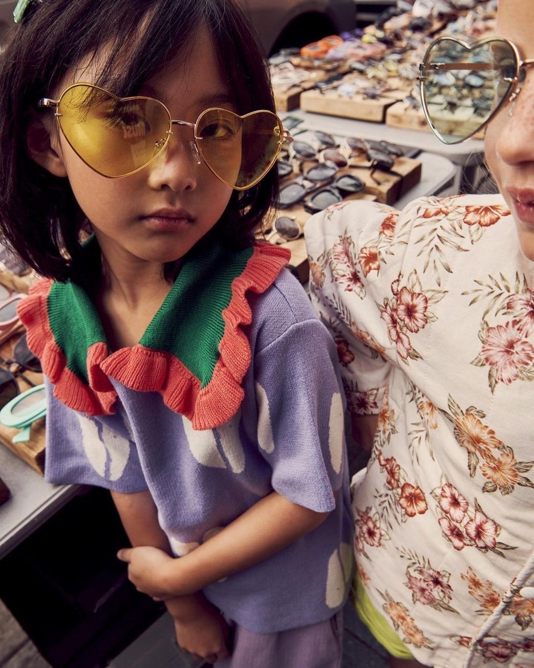 Two young girls with heart-shaped sunglasses pose outdoors among sunglasses displays; one wears a floral top, the other a ruffled sweater.