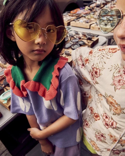 Two young girls with heart-shaped sunglasses pose outdoors among sunglasses displays; one wears a floral top, the other a ruffled sweater.