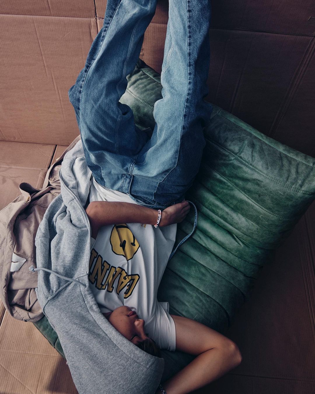 Person lying upside down on a green cushion inside a cardboard structure, wearing jeans, t-shirt, and grey zip-up hoodie, casual relaxed style.