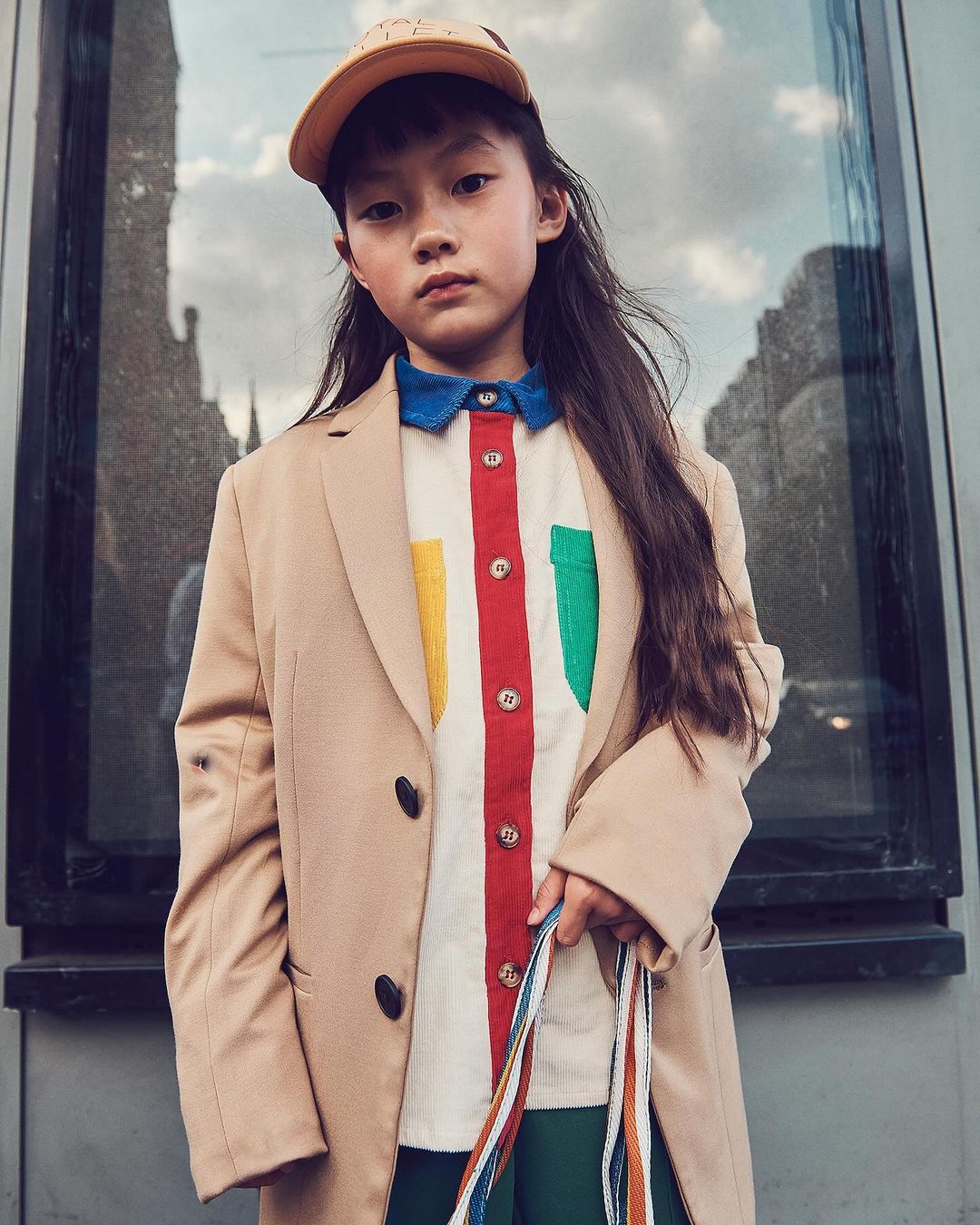 Stylish Asian child with a tan cap and multicolored shirt wearing a tan jacket in an urban setting, fashionably posing and looking at camera.