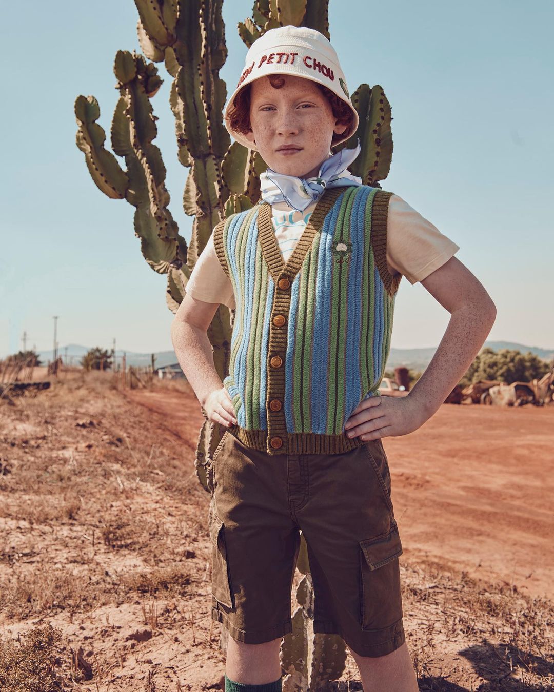 A young, fair-skinned child poses outdoors with a cactus, wearing a colorful vest, bucket hat, and shorts in warm, natural light.