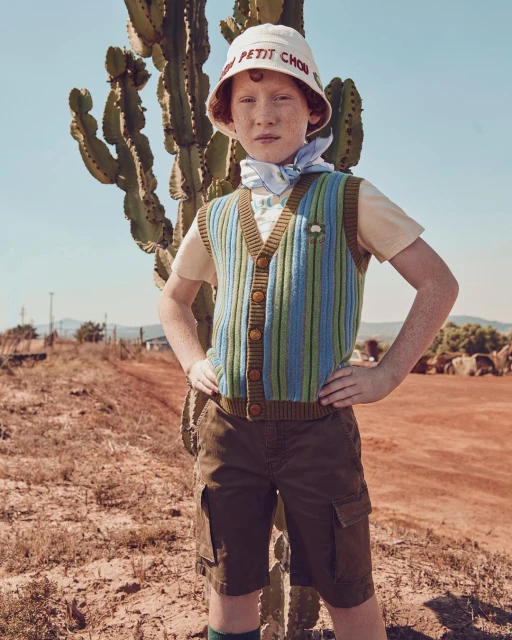 A young, fair-skinned child poses outdoors with a cactus, wearing a colorful vest, bucket hat, and shorts in warm, natural light.