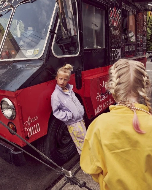 Two blond girls in front of a red food truck with black trim, casual jackets, and braided hair, shot during daylight hours outdoors.