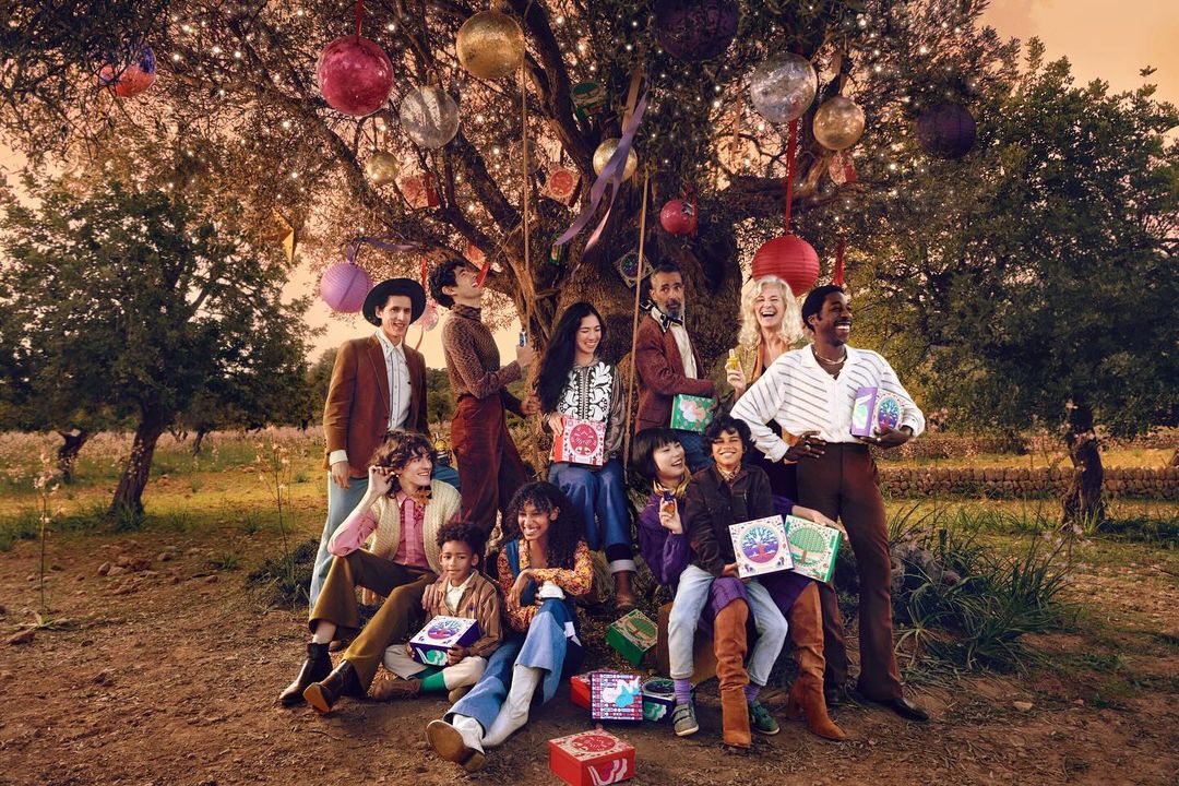 Group of diverse people gathered under a tree decorated with colorful ornaments and lights, holding boxes, outdoors in natural setting.