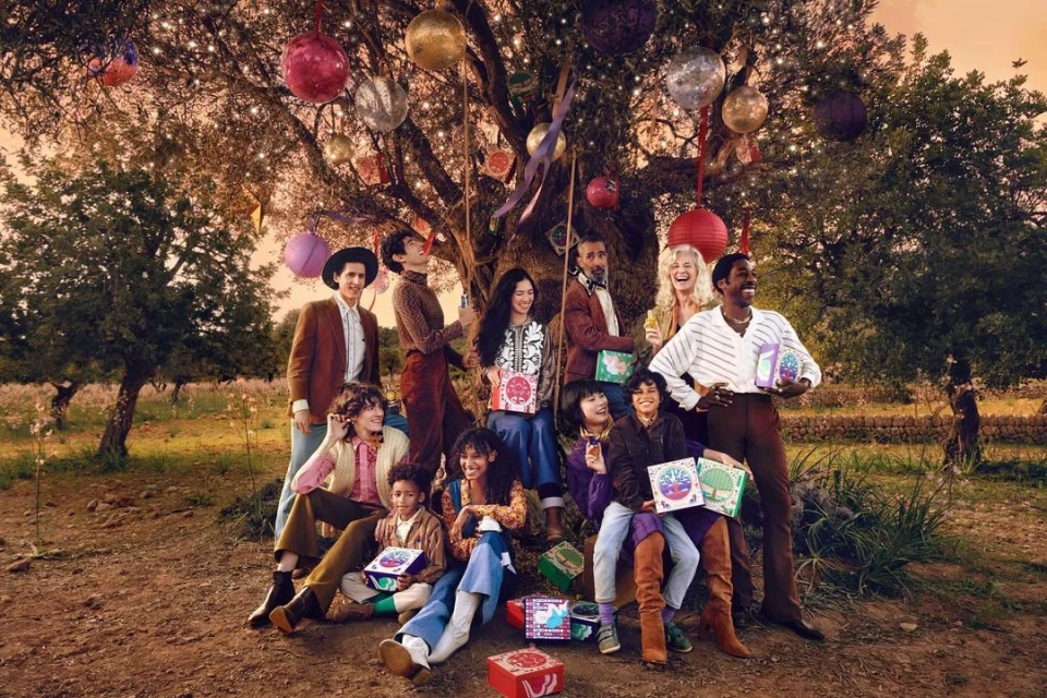 Group of diverse people gathered under a tree decorated with colorful ornaments and lights, holding boxes, outdoors in natural setting.