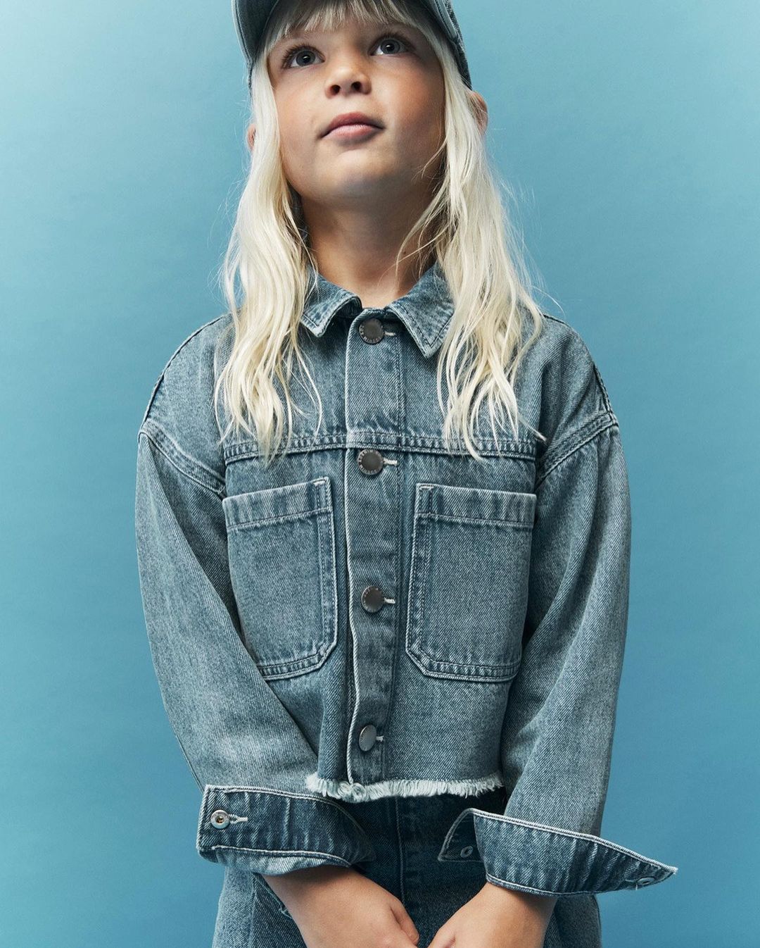 Young child model wearing a denim jacket and hat against a blue backdrop, looking up in a fashion or lifestyle shot.