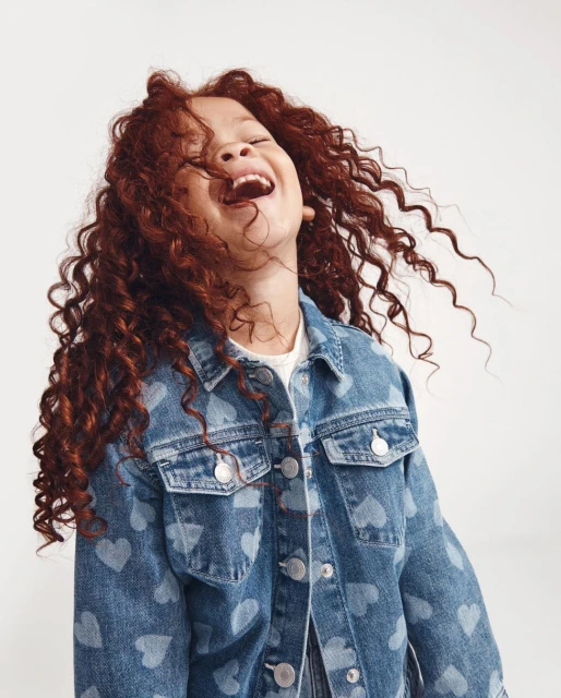A joyful young girl with curly red hair is laughing and wearing a denim jacket with heart pattern against a neutral background.