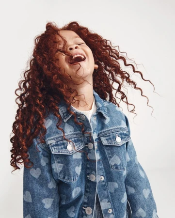 A joyful young girl with curly red hair is laughing and wearing a denim jacket with heart pattern against a neutral background.