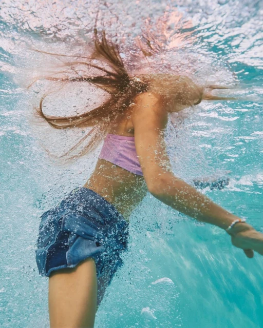 Underwater view of a young woman with long hair wearing a bikini top and denim skirt, partially submerged in blue water with bubbles.