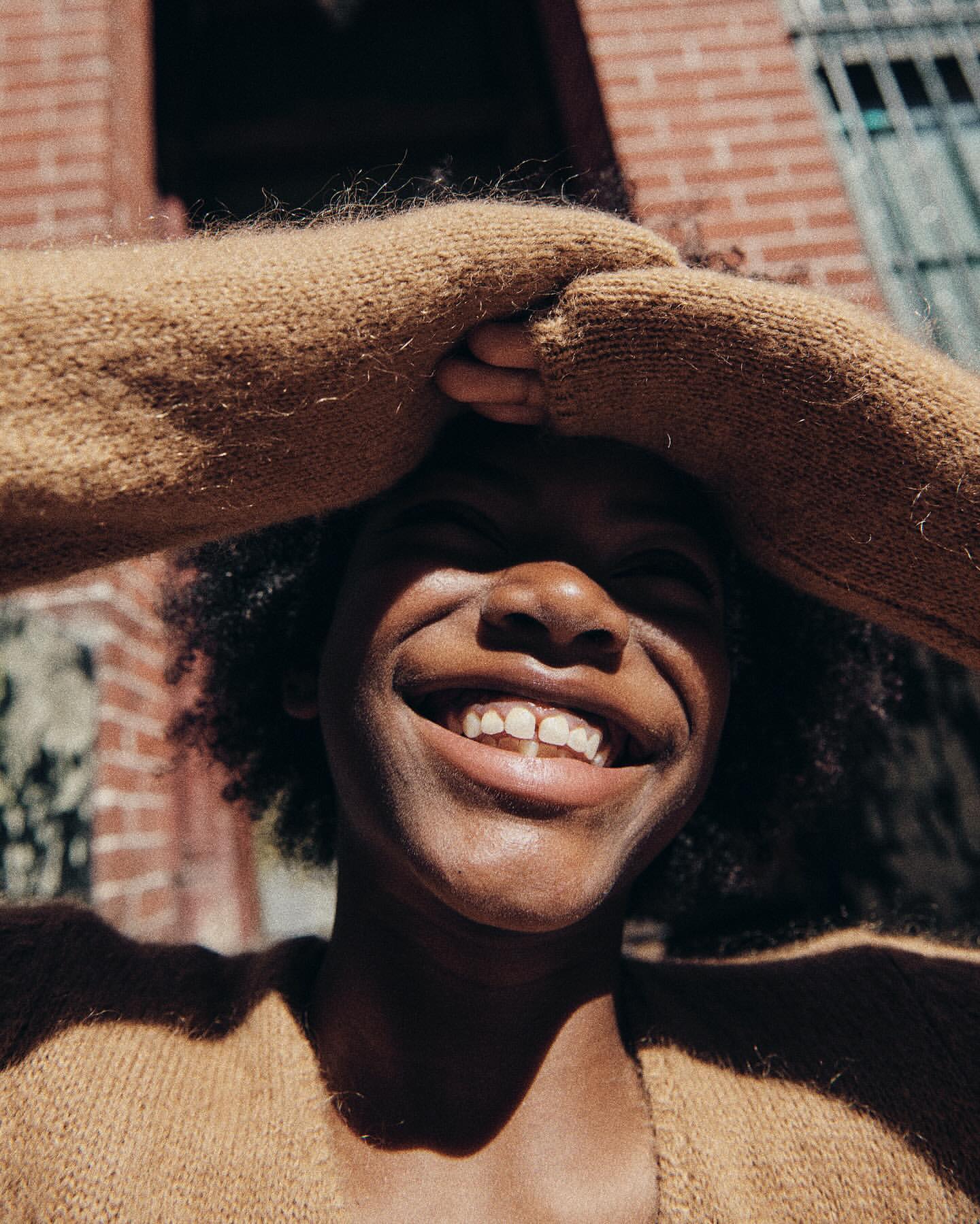 Close-up of a smiling African-American woman with her hands on her head, wearing a tan sweater, with a brick building in the background.