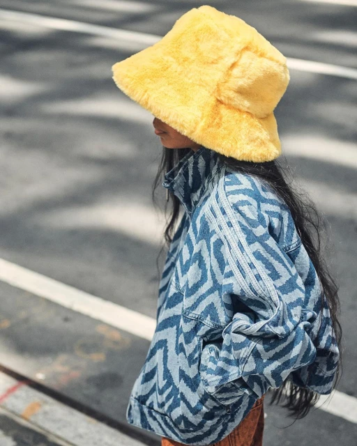 A girl wears a patterned denim jacket and a yellow fuzzy bucket hat on city street, showcasing fashion style and urban lifestyle.