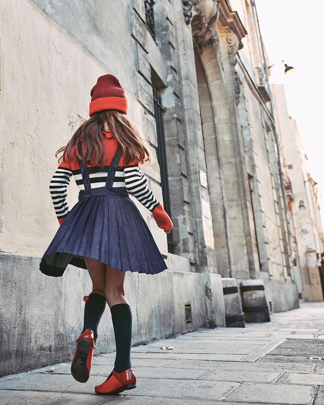 A girl in a stylish navy dress, a red-and-white striped sweater, and a red hat walks down a stone street near a building.