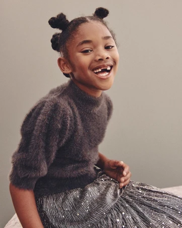 Young Black girl smiling with hair in multiple buns wearing a dark fuzzy sweater and shiny metallic skirt against grey