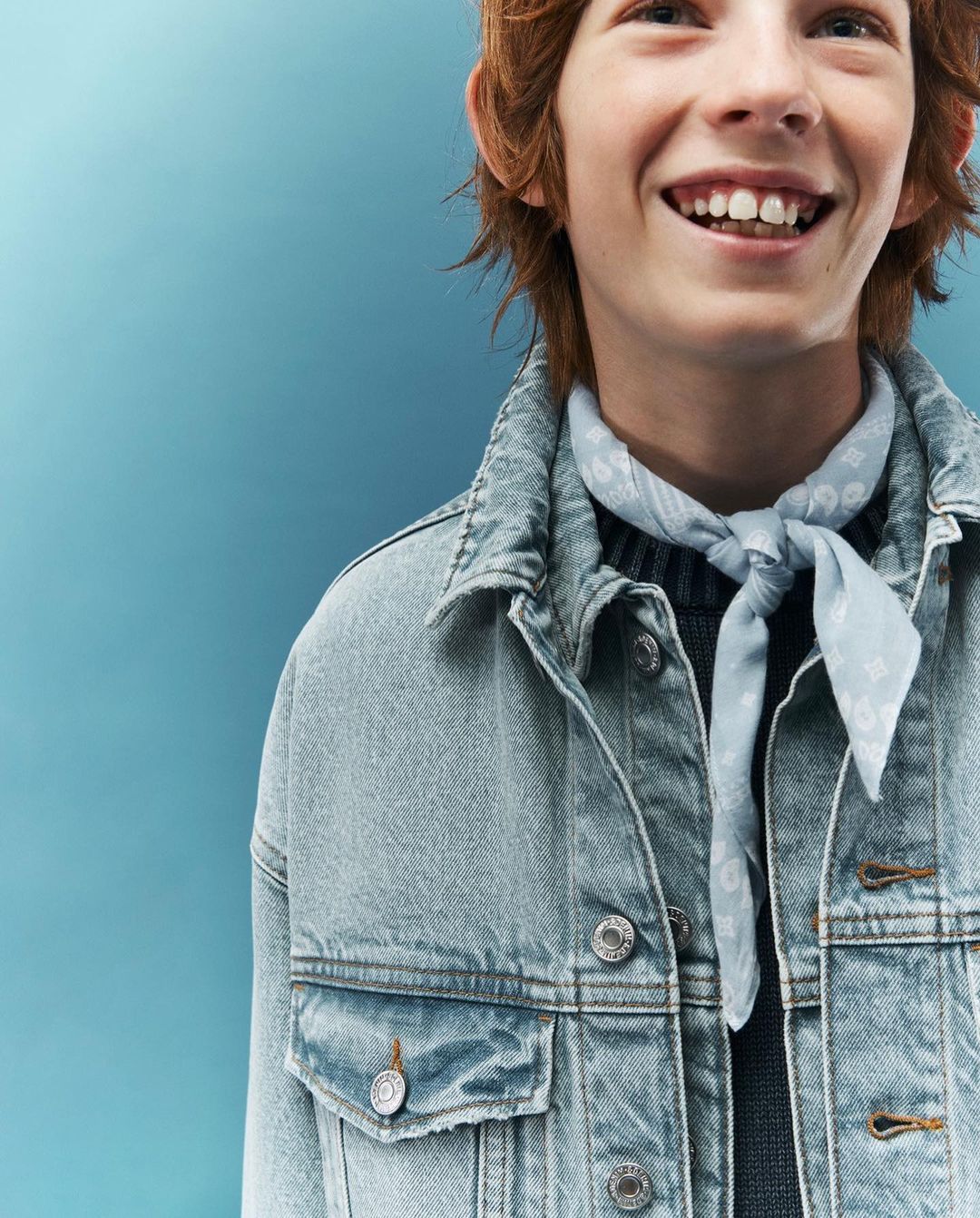 Smiling boy with red hair wears a denim jacket and patterned kerchief in a studio shot against a solid light blue background.