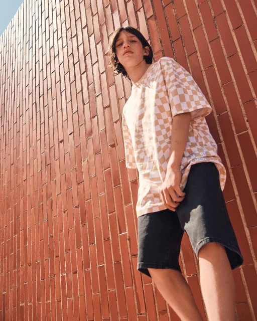 Young adult wearing a checkered t-shirt and shorts poses against a brick wall in an outdoor setting during daylight.