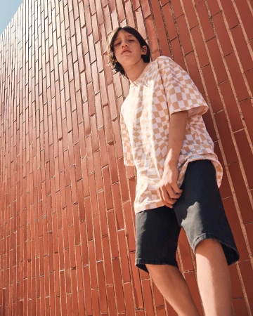 Young adult wearing a checkered t-shirt and shorts poses against a brick wall in an outdoor setting during daylight.