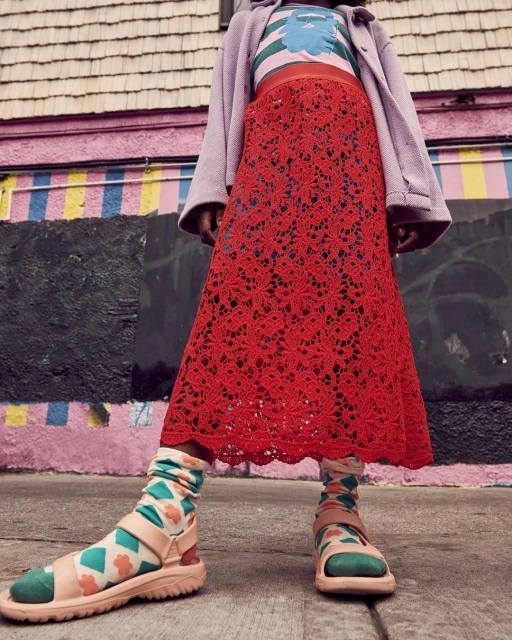 Person wearing a red, floral lace skirt, printed socks and strappy sandals standing in front of a pink building with stripes.