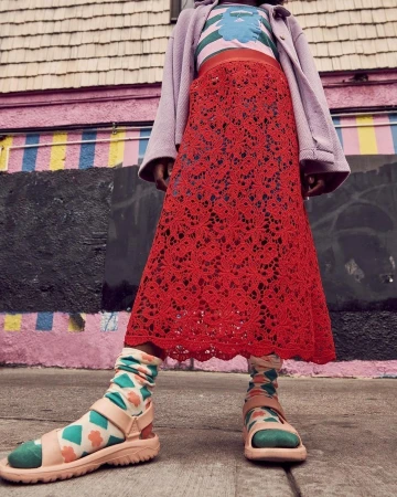 Person wearing a red, floral lace skirt, printed socks and strappy sandals standing in front of a pink building with stripes.