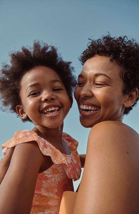 Close up shot of an African-American mother and daughter laughing outdoors in bright sunlight with a clear blue sky background.