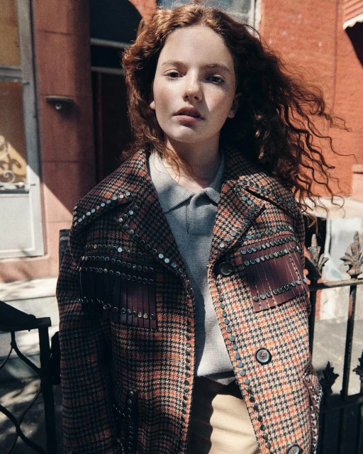 An outdoor portrait shot of a young red-haired woman wearing a houndstooth coat with embellishments, standing near a brick building.