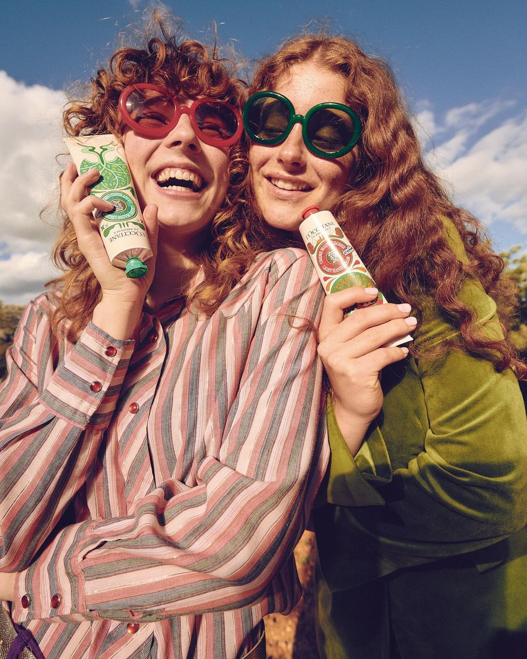 Two happy women with red curly hair wear 70s-style glasses, holding hand cream tubes against a blue sky with soft clouds and trees.