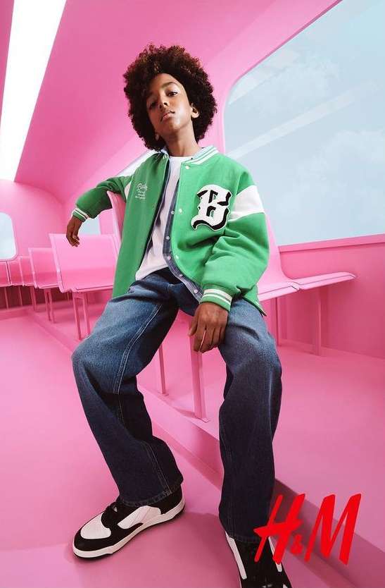 Young boy sits in a pink train car wearing a green and white letterman jacket, blue jeans, and black and white sneakers, H&M logo in corner.
