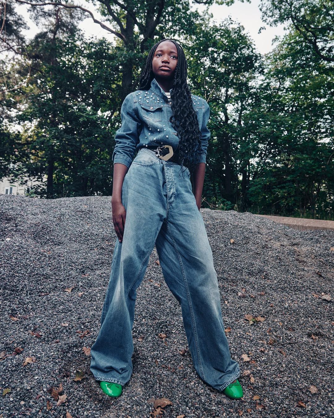 A young Black woman stands on a gravel slope wearing a denim shirt and oversized jeans in an outdoor wooded environment during daytime.