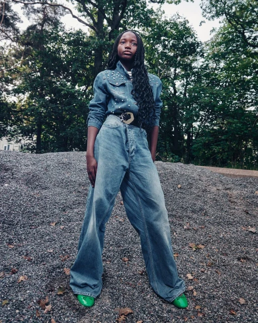 A young Black woman stands on a gravel slope wearing a denim shirt and oversized jeans in an outdoor wooded environment during daytime.