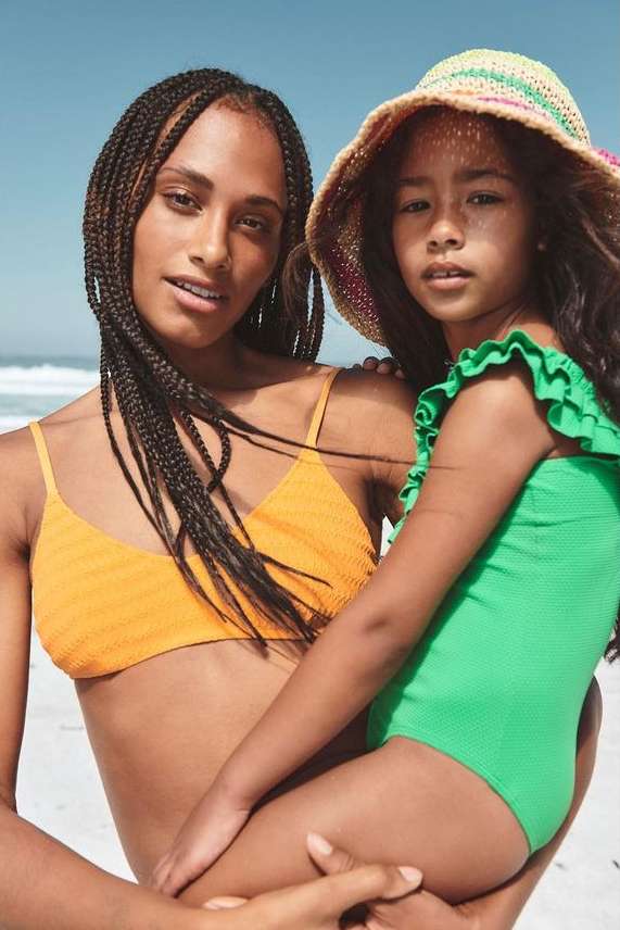 Woman and child in swimwear on beach. Adult female in orange bikini holds young girl in green swimsuit with straw hat.