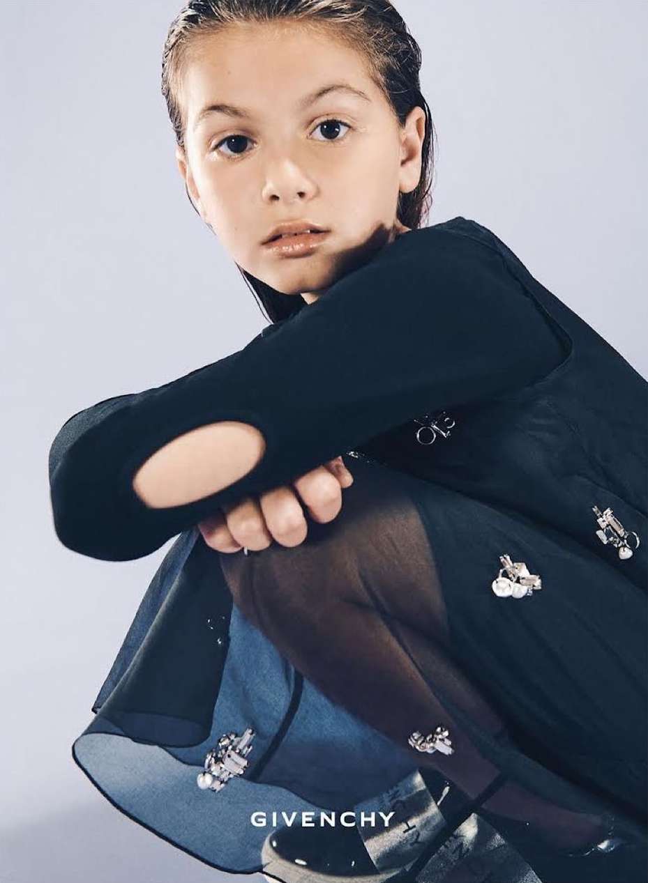 Child crouching in a dark dress with metallic embellishments against a neutral backdrop, photographed by Franck Malthiery.