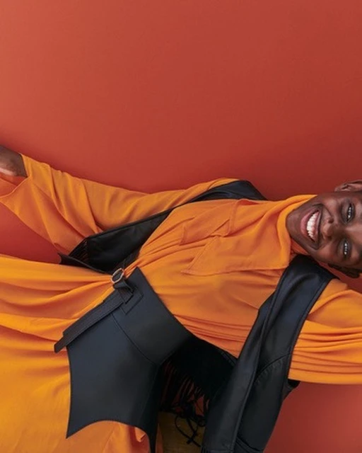 Fashionable adult in an orange top with a black vest smiles against a solid orange background, captured in a studio portrait.