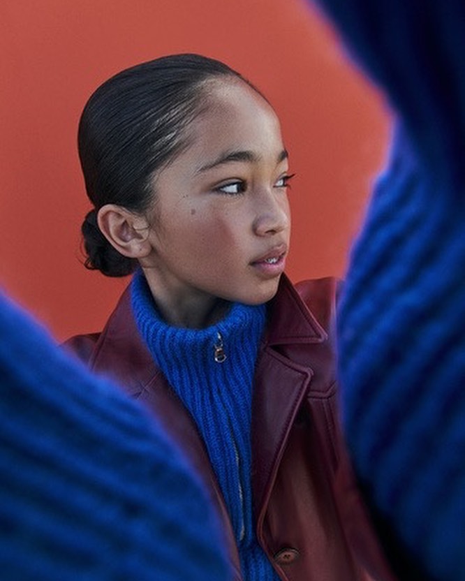 Portrait of a young person with slicked-back hair and a burgundy jacket over a blue turtleneck against an orange backdrop.