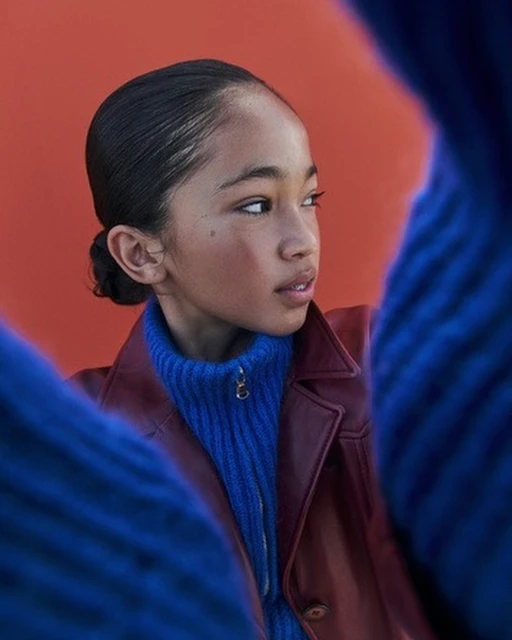 Portrait of a young person with slicked-back hair and a burgundy jacket over a blue turtleneck against an orange backdrop.