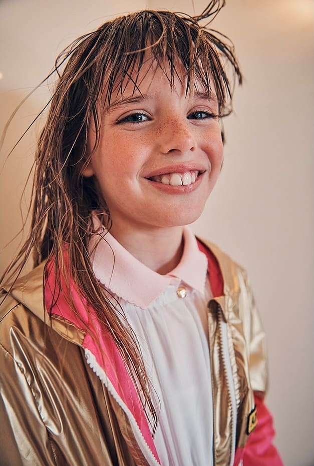 Smiling child with wet brown hair and freckles, wearing a gold jacket with pink accents. Head and shoulders portrait against a plain wall.