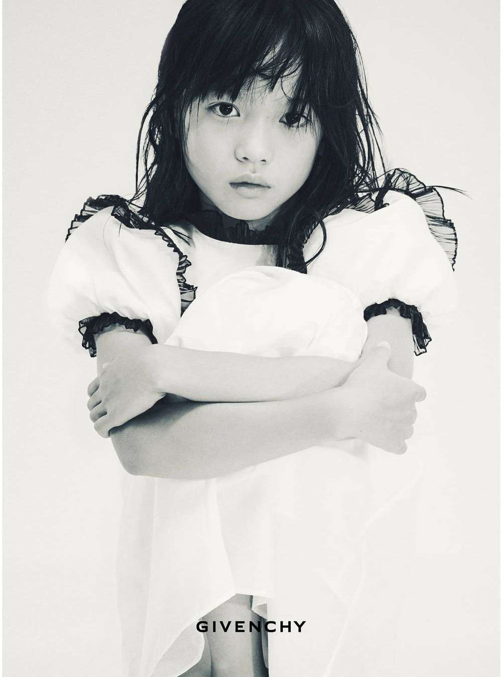 Young girl in a white dress with black trim, arms crossed, staring at the camera in a black and white indoor fashion shot.