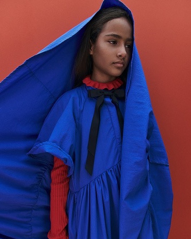 Portrait of a young girl with brown hair, wearing a blue dress with a hood and black bow, against a red studio background.