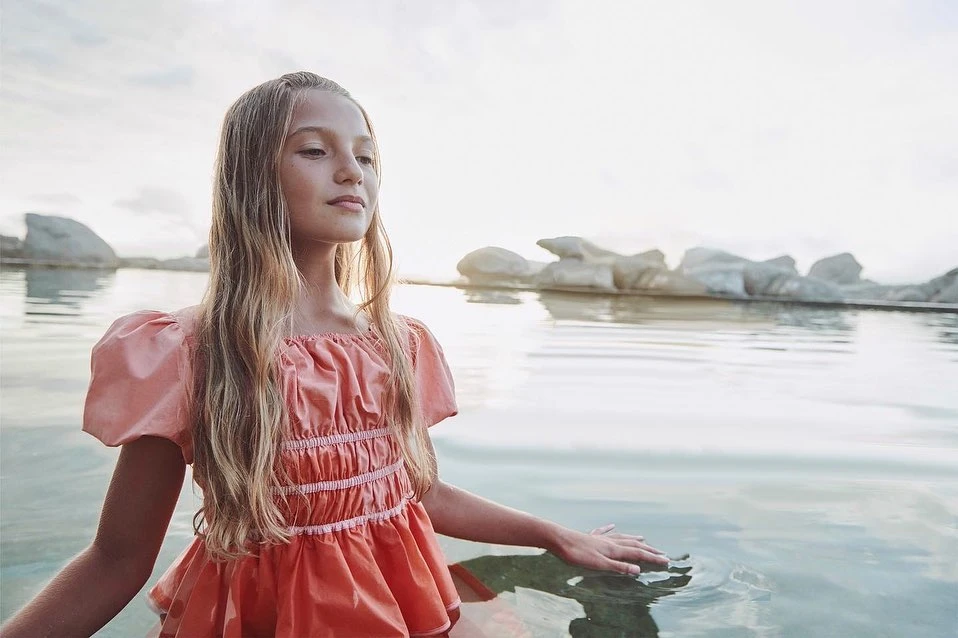 Girl in peach dress stands in water with distant rock formations, creating a serene and ethereal outdoor portrait.