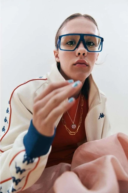 Portrait of a fair-skinned young woman wearing blue glasses, a Kappa track jacket, jewelry, and with light blue nail polish in a studio setting.