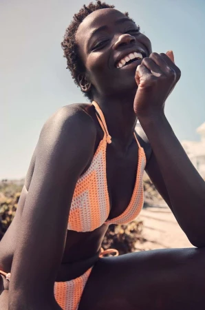 Smiling black woman wearing a peach and white patterned crochet bikini outdoors under bright sunlight.