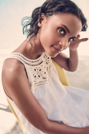 Close-up portrait of a woman in a white crochet-trimmed top looking back over her shoulder while seated on a yellow chair.