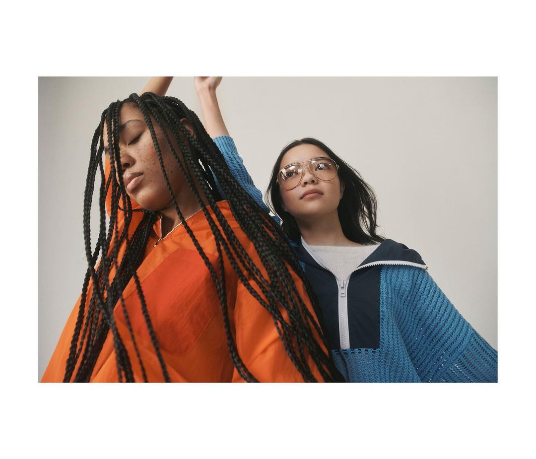 Two women in an indoor setting, one with braids and an orange top and the other with glasses and a navy and teal sweater.