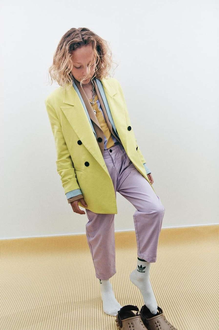 Young adult woman in a yellow blazer and light purple pants standing indoors with her head down, wearing white socks and brown shoes.