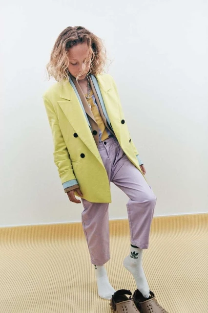 Young adult woman in a yellow blazer and light purple pants standing indoors with her head down, wearing white socks and brown shoes.