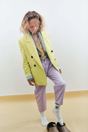 Young adult woman in a yellow blazer and light purple pants standing indoors with her head down, wearing white socks and brown shoes.