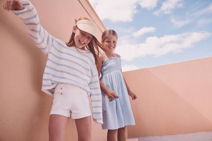 Two young girls smiling outdoors against a curved peach wall under a partly cloudy blue sky